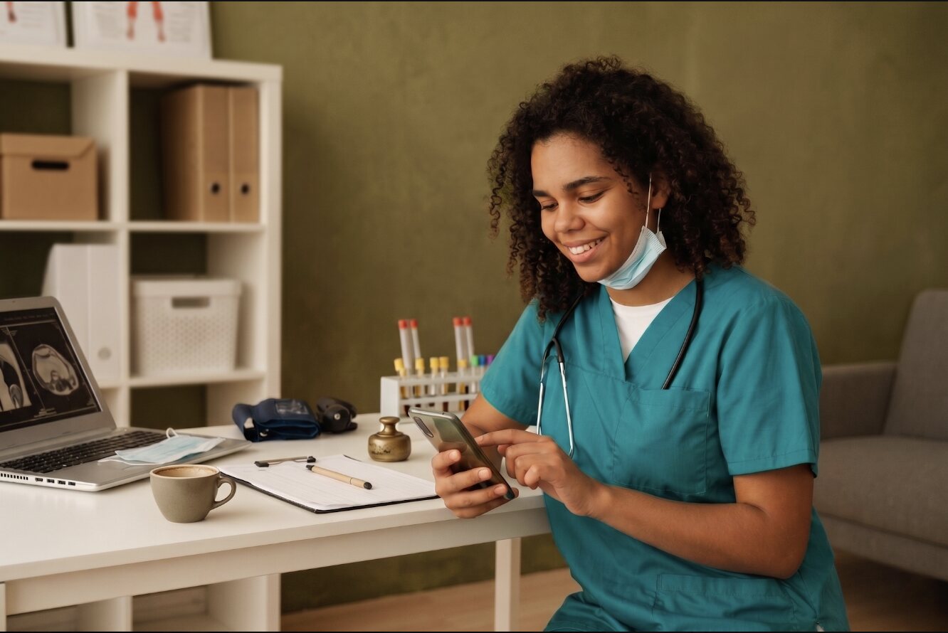 A smiling nurse in teal scrubs checks her phone at a clinic desk, with test tubes and a blood pressure cuff beside her.