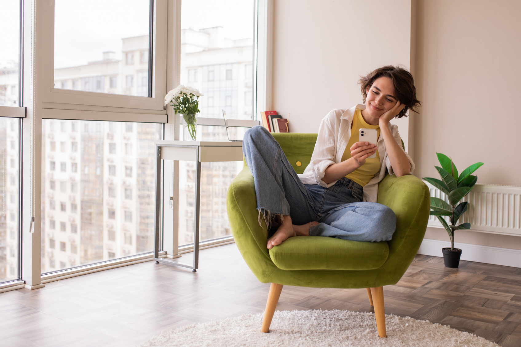 A woman curled up in a green armchair by a sunny window smiles at her phone.