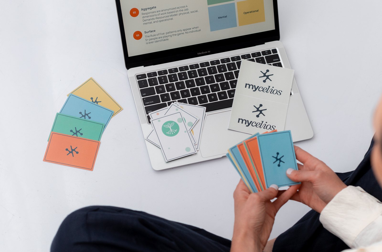 Hands hold a fan of colorful Mycelios cards beside a laptop showing the Latitude game, with Mycelios-branded cards and a sleeve on the table.