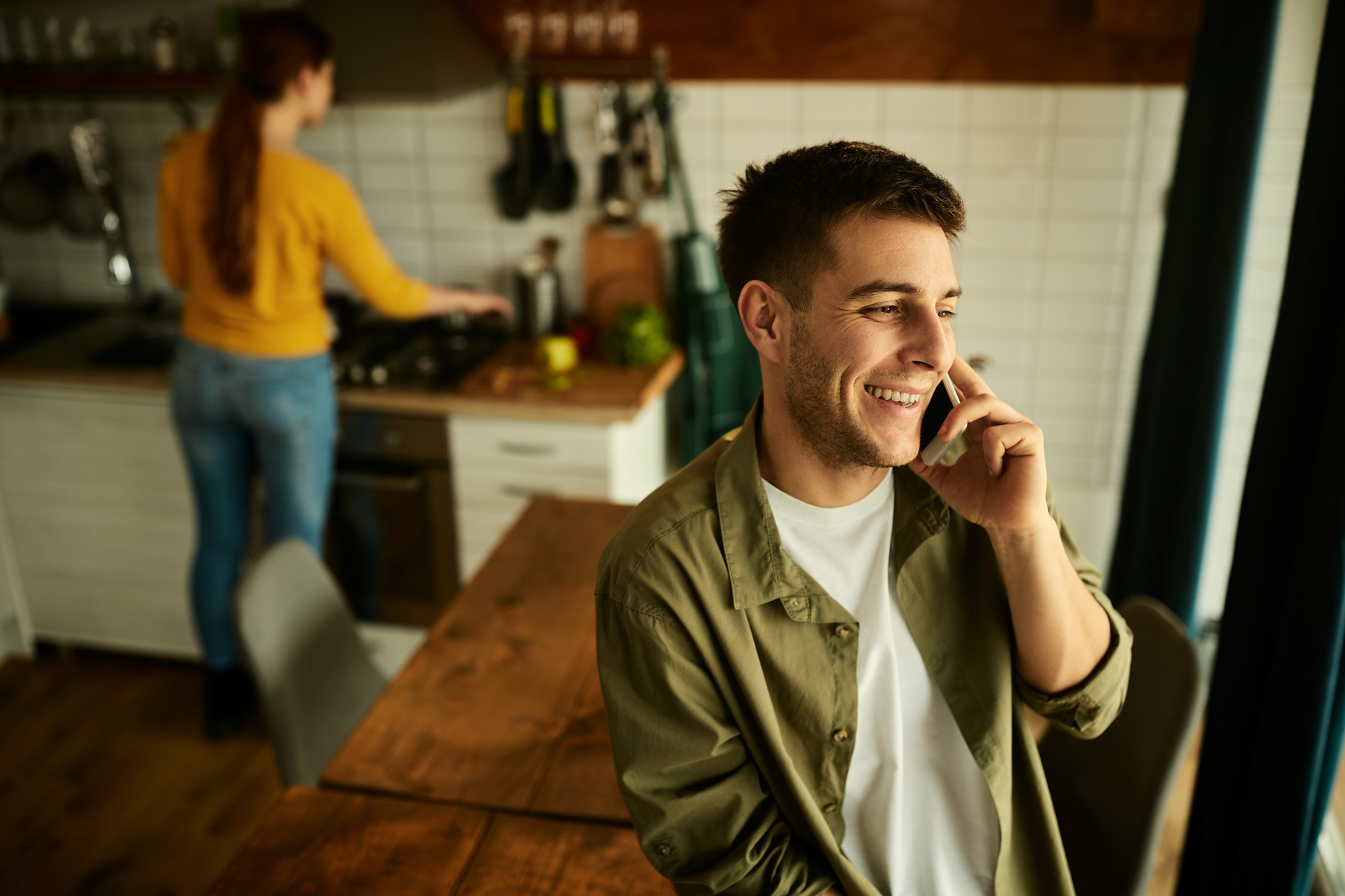 A smiling man talks on the phone at a kitchen table while someone cooks in the background.