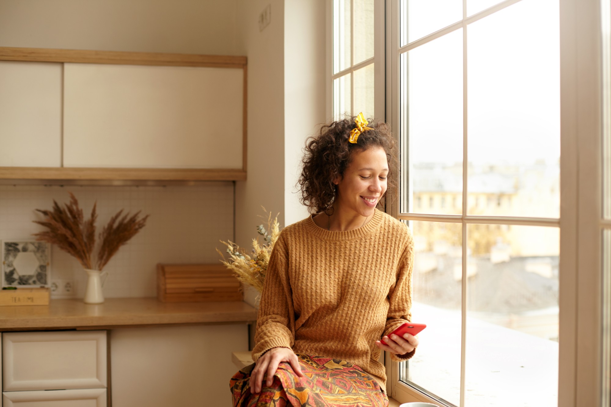 A smiling woman in a mustard sweater sits by a tall window at home, looking down at her phone.